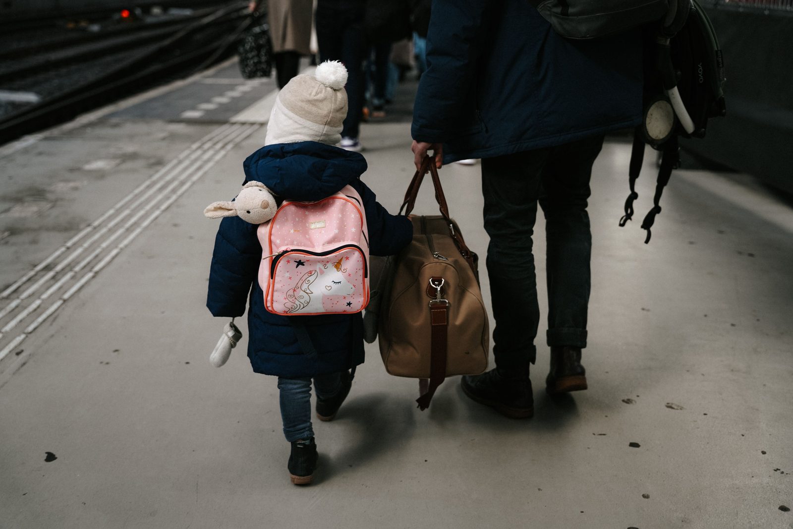 A child carrying a backpack walks along a station platform beside a parent carrying luggage